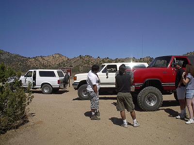 1990 Ford Bronco - group of people gather around a white SUV and a lifted red SUV parked on a dirt clearing among boulders.
