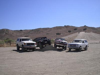 1990 Ford Bronco - Four 4x4 vehicles, white and red, parked side by side facing forward in a dusty desert clearing below dry hills.
