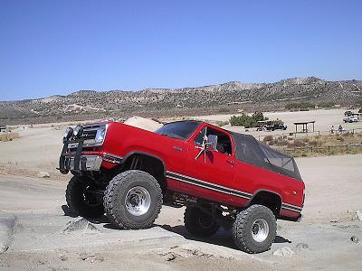 1990 Ford Bronco - red lifted SUV resembling a Dodge Ramcharger on oversized tires sits in a sandy desert lot below dry hills.