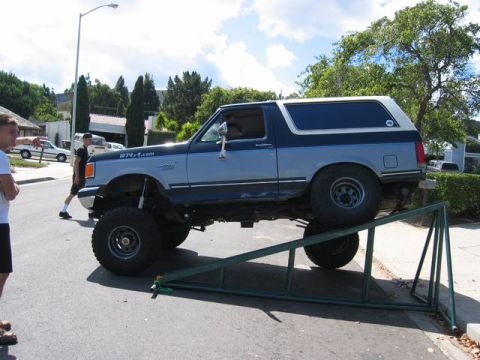1989 Ford Bronco - A lifted Ford Bronco is positioned on metal ramps, with two people observing nearby.