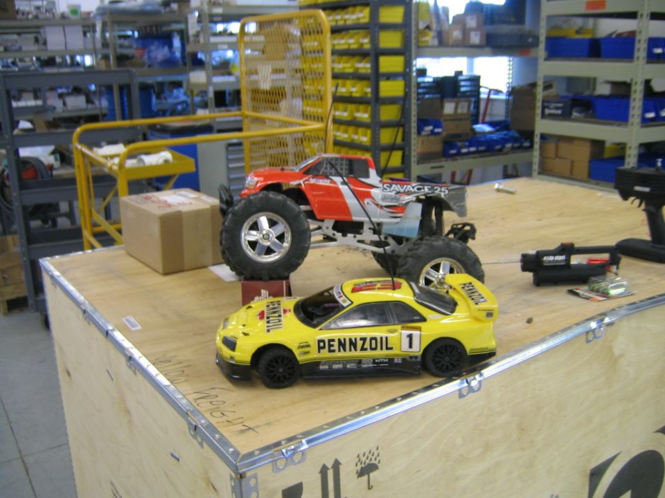 Two RC cars, a yellow Pennzoil-liveried sports car and a red and silver monster truck labeled Savage 25, sit on a wooden surface in a warehouse