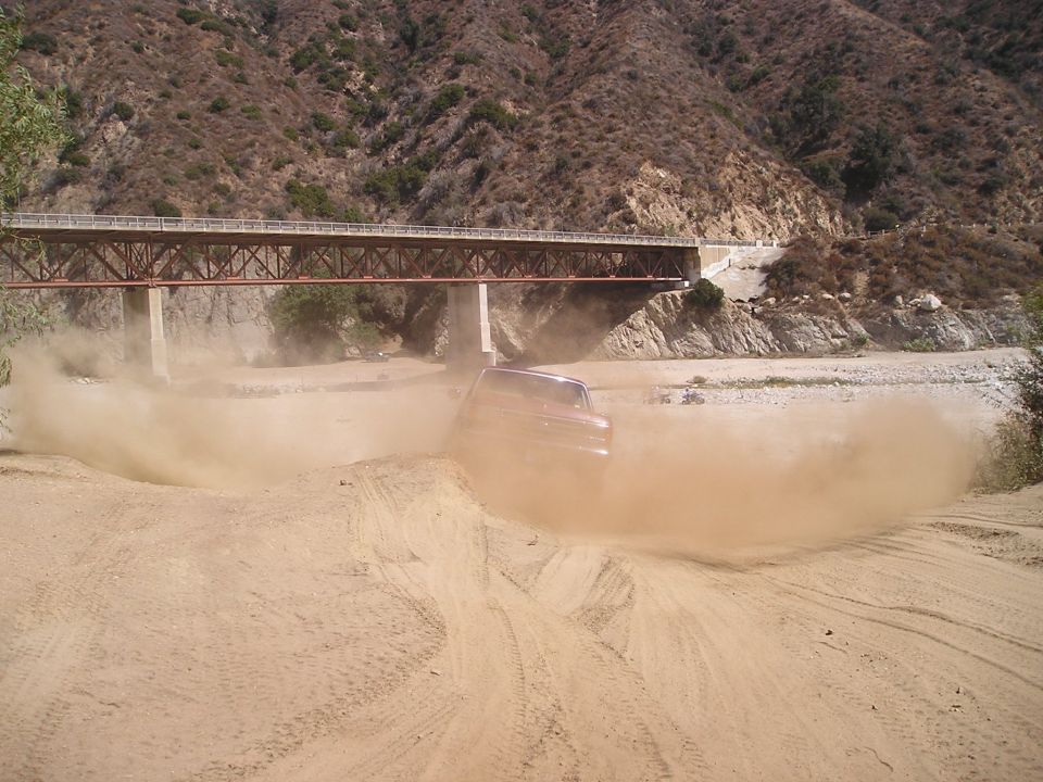 Dark red vehicle kicking up a huge dust cloud in a dry wash under a truss bridge