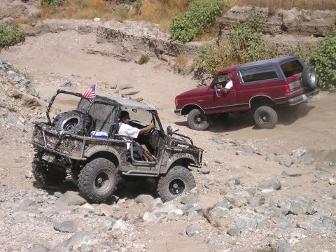 1989 Ford Bronco - A red 1987 Ford Bronco navigates a challenging off-road trail, with another modified vehicle in the foreground.
