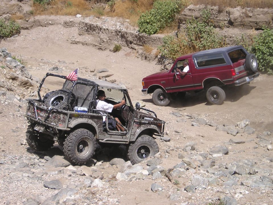 Red 1987 Ford Bronco climbing a rocky dirt hill, with another off-road vehicle visible