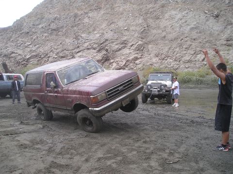 1989 Ford Bronco - A maroon Ford Bronco is stuck in the mud with its front wheel lifted, while another vehicle waits behind it.
