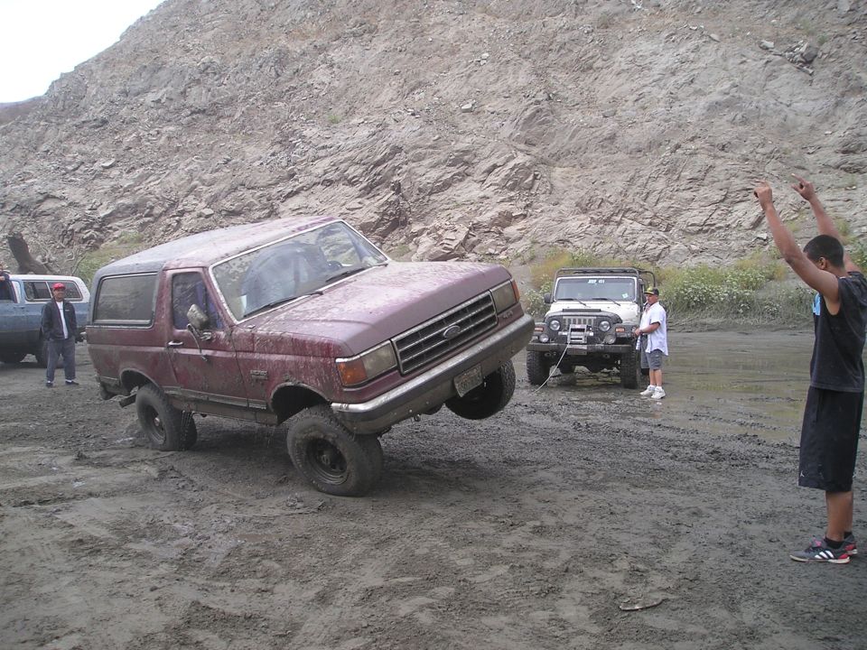 Maroon Ford Bronco with front wheel lifted off the ground, stuck in mud