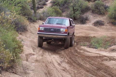 1992 Ford Bronco - This maroon Ford Bronco is navigating a sandy, off-road trail with desert vegetation surrounding it.