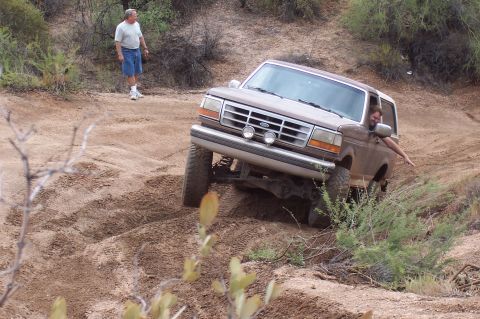1992 Ford Bronco - A 1992 Ford Bronco is shown off-roading on a sandy, uneven terrain with a person visible in the driver's seat.