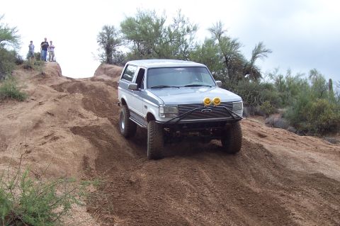 1992 Ford Bronco - Silver-grey lifted Ford Bronco flexing over a dirt berm on an off-road trail, fitted with a tube front bumper, winch and two yellow smiley-…
