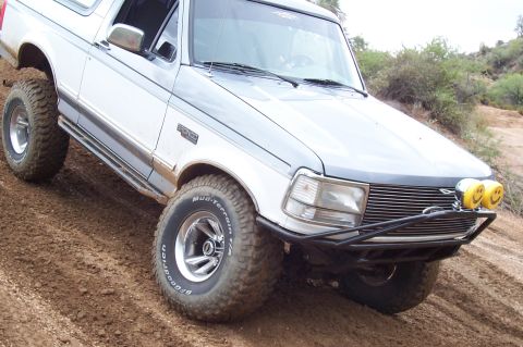 1992 Ford Bronco - A silver/light gray Ford Bronco full-size SUV shown from a front three-quarter angle, climbing dirt terrain with BFGoodrich Mud-Terrain T/A…