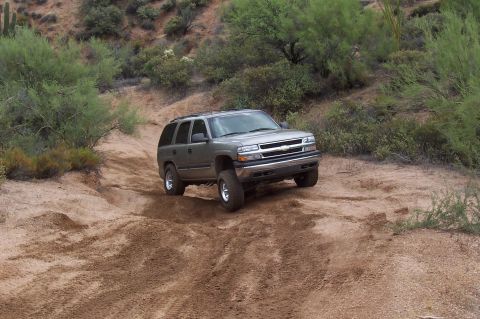 1992 Ford Bronco - Chevrolet Tahoe navigates a sandy off-road path with desert vegetation in the background.