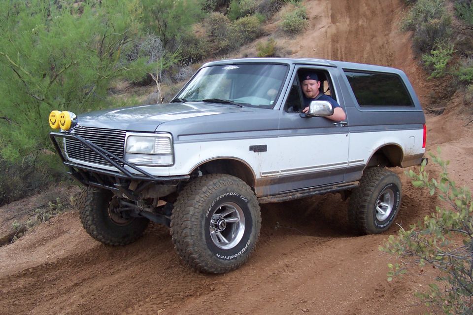 Man driving a 1992 Ford Bronco on a dirt trail with aftermarket bumper and lights