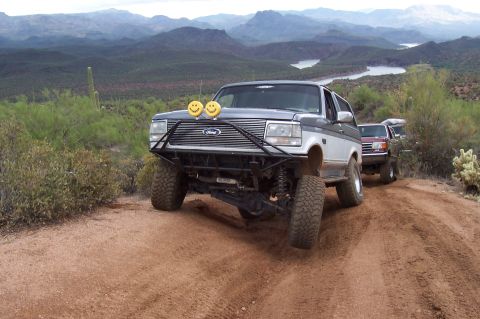 1992 Ford Bronco - A modified Ford Bronco navigates a dusty trail in a mountainous desert landscape with a lake in the background.