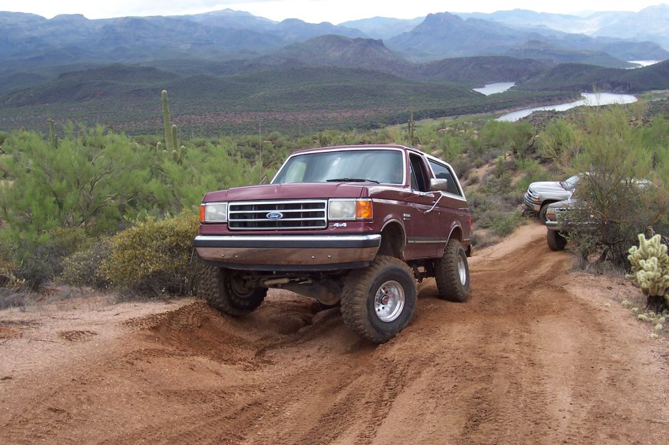 Maroon Ford Bronco with lifted suspension driving on a dirt road in a desert landscape
