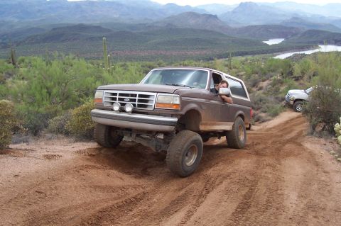 1992 Ford Bronco - Brown 1992 Ford Bronco navigates a dusty off-road trail with a scenic desert backdrop and another vehicle in the distance.