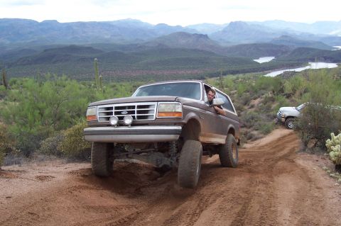 1992 Ford Bronco - A man drives a lifted Ford Bronco on a dusty trail in a desert landscape with mountains and a lake visible.