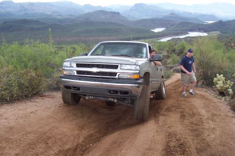 1992 Ford Bronco - A silver pickup truck drives on a dusty mountain road with a man standing nearby.