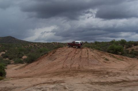 1992 Ford Bronco - A red SUV with a white camper shell sits atop a dusty hill under a dramatic, cloudy sky.