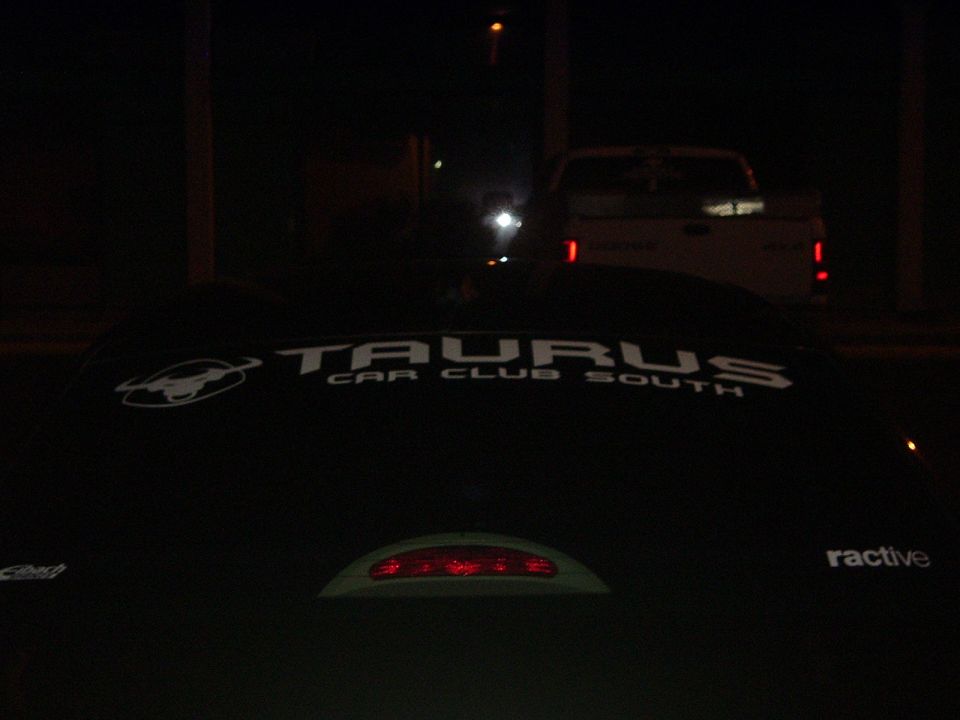 Car with 'TAURUS CAR CLUB SOUTH' decal on the rear window, another truck is visible in the background