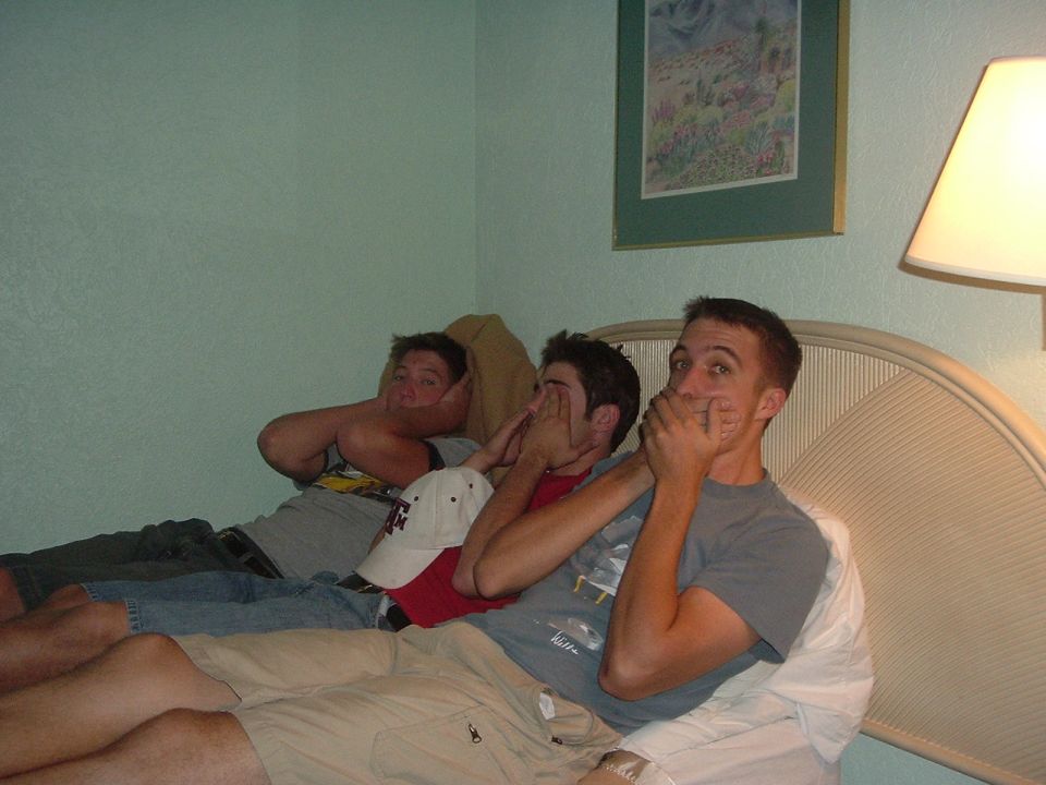Three men on a bed covering their mouths, with a Supermotors logo in the corner