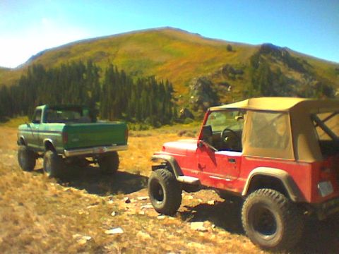 1979 Ford Bronco - Two off-road vehicles, a green Ford pickup and a red Jeep, are parked on a dirt track with a mountain landscape behind them.