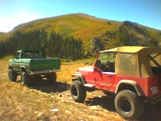 Green 1970s Ford pickup truck and red Jeep Wrangler parked on a dirt road with a mountain in the background