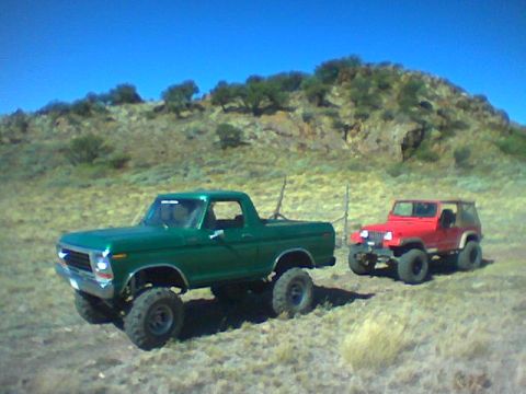 1979 Ford Bronco - Two off-road vehicles, a green Ford pickup and a red Jeep, are parked in a dry, hilly landscape under a clear blue sky.