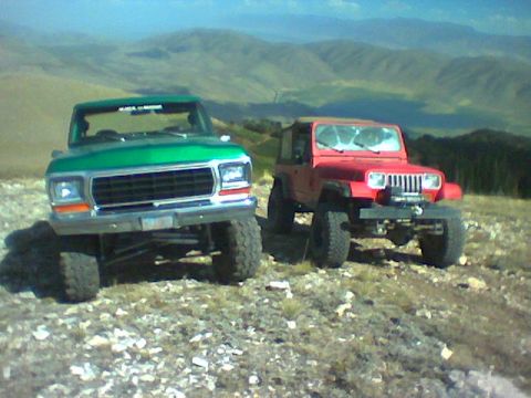 1979 Ford Bronco - Two off-road vehicles, a green 1979 Ford truck and a red Jeep, are parked on a rocky mountain slope under a clear sky.