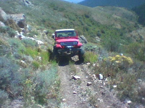 1979 Ford Bronco - Red Jeep Wrangler navigates a rough dirt path through a mountainous, brush-covered landscape.