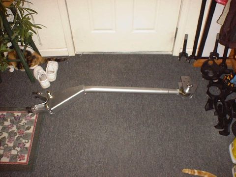 1988 Ford Bronco - Full-length view of a silver-painted custom radius arm laid out on gray carpet, with a boxed triangular plate section and dual axle-mount c…