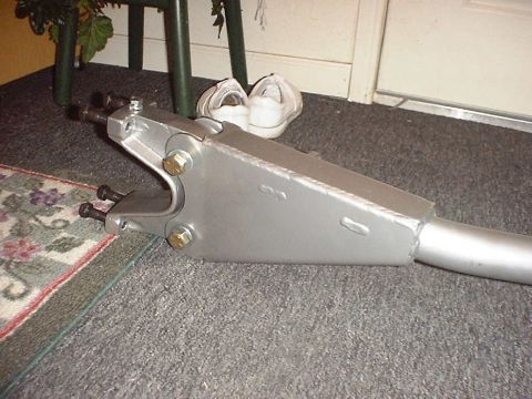 1988 Ford Bronco - A freshly painted silver fabricated suspension link or radius arm lying on gray carpet indoors, showing boxed plate construction with visib…