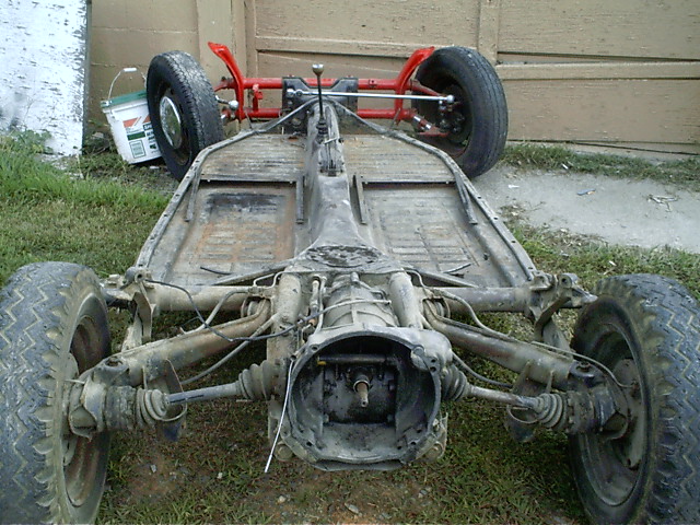 Stripped VW Beetle floor pan chassis on grass, rear view showing suspension and red front beam, no engine installed