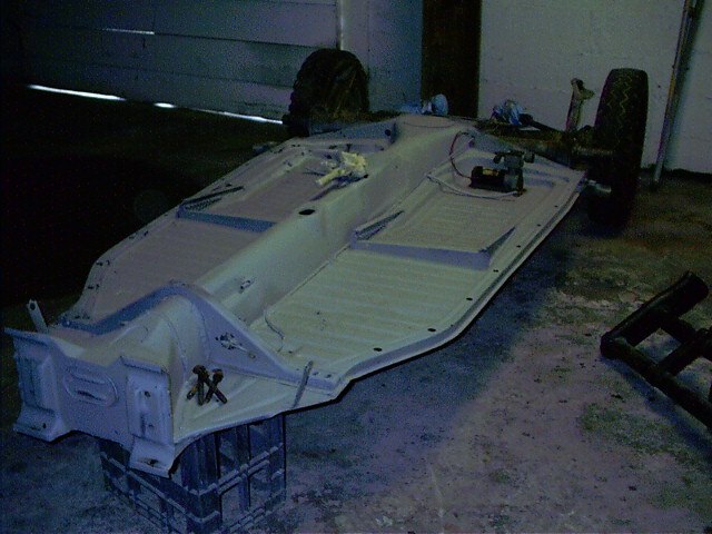 Stripped VW Beetle floor pan chassis in gray primer on blocks in a garage, rear wheels attached