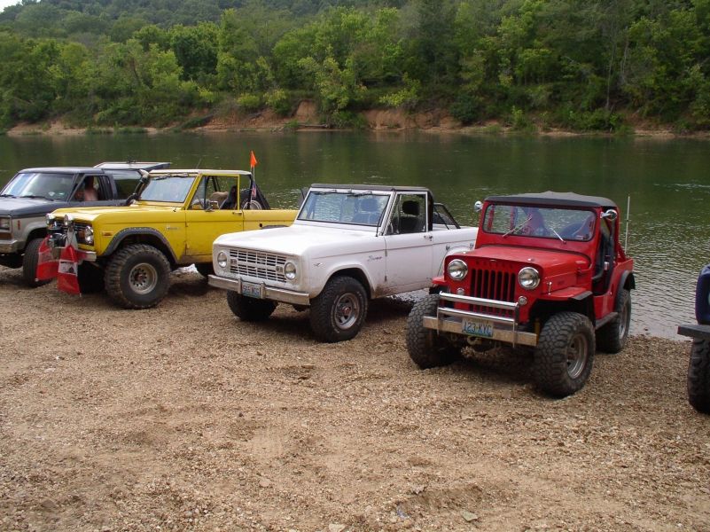 Several off-road vehicles parked on a gravel shore next to a river. A white Ford Bronco is in the foreground