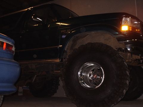 1996 Ford Bronco - A dark green Ford Bronco with oversized tires is parked indoors next to a blue vehicle.