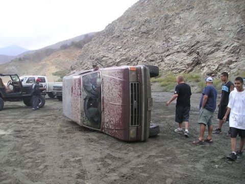 1990 Ford Bronco - maroon full-size SUV lies tipped onto its side on a gravel lot while a group of young men gather around it beneath a rocky cut hillside.