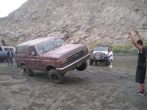 1990 Ford Bronco - mud-covered maroon Ford Bronco crawls through deep sandy ruts, lifting a front tire, as a bystander throws his arms up beside a Jeep.