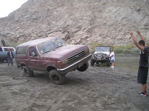 Muddy maroon Ford Bronco lifting a front wheel in deep ruts as a person cheers nearby