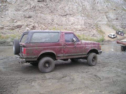1990 Ford Bronco - lifted maroon Ford Bronco coated in dried mud sits in profile on a dirt lot, sporting a rear-mounted spare tire and a quarry wall behind.
