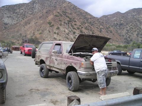 1990 Ford Bronco - man bends over the open engine bay of a mud-splattered lifted maroon Ford Bronco parked in a dirt lot below dry mountains.