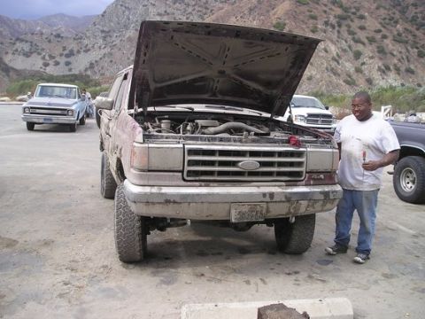 1990 Ford Bronco - man stands at the front of a lifted maroon Ford with its hood raised, exposing the engine, in a dirt lot framed by mountains.