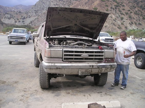 Man standing beside a maroon Ford truck with its hood up showing the engine in a dirt lot
