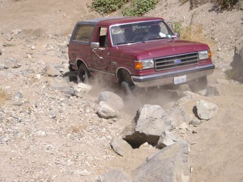 1990 Ford Bronco - maroon Ford Bronco kicks up dust as it climbs over a field of large rocks on a dusty off-road trail.