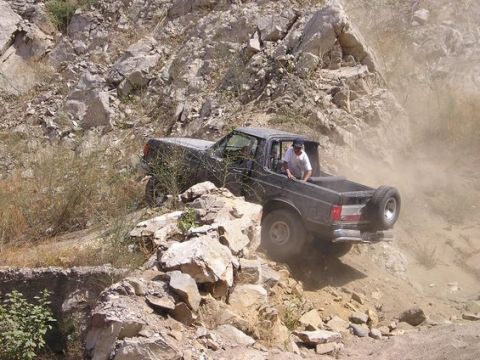 1990 Ford Bronco - dark single-cab pickup truck claws its way up a steep rocky shelf, kicking up dust, with a man standing in the open bed.