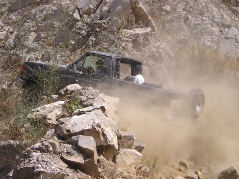 1990 Ford Bronco - dark pickup truck strains up a steep rocky ledge, nearly engulfed in a billowing cloud of dust, with a person in the bed.