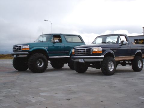 1996 Ford Bronco - Two lifted Ford Broncos, a teal 1996 model and a navy blue one, are parked outdoors under an overcast sky.