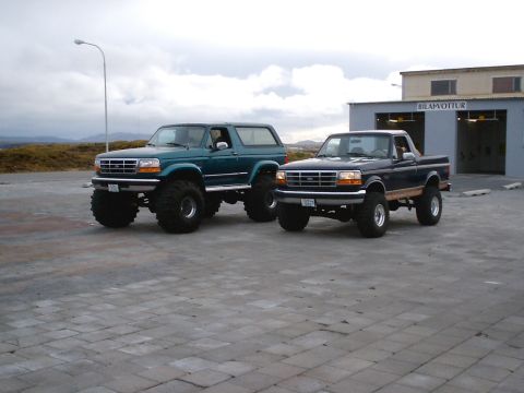 1996 Ford Bronco - Two lifted Ford trucks, a Bronco and a pickup, are parked outside a building with "BILAPVOTTUR" signage.