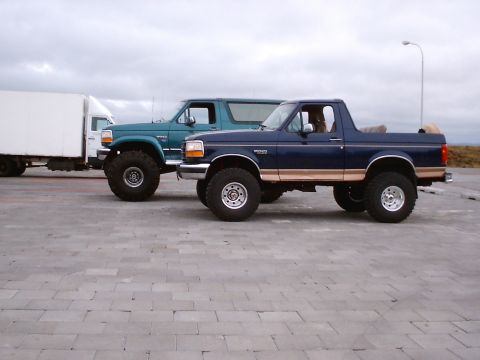 1996 Ford Bronco - Two Ford Broncos are parked side-by-side on a paved lot under an overcast sky.