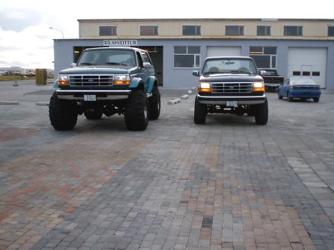 1996 Ford Bronco - Two lifted Ford Broncos, a teal one and a dark blue one, are parked side-by-side in front of a building.