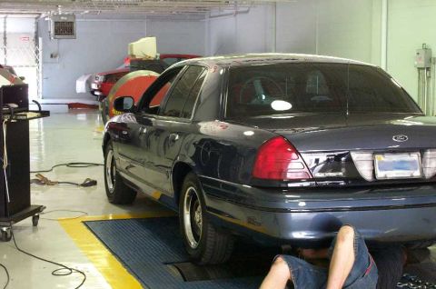 1998 Ford Crown Victoria - A mechanic works on the underside of a dark sedan while another vehicle is parked behind it in a garage.